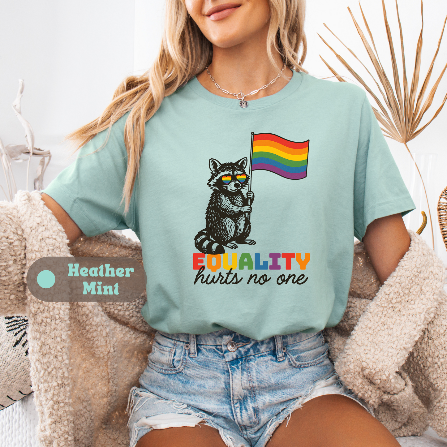 A woman wearing a Heather Mint t-shirt with a raccoon holding a rainbow flag and the phrase βEQUALITY hurts no one.β A tag marks the shirt as Heather Mint.
