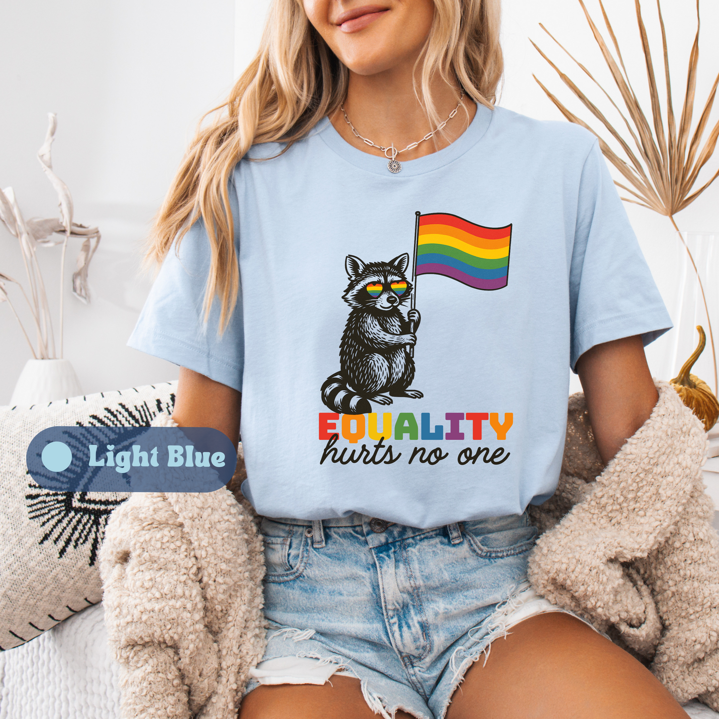 A woman wearing a Light Blue t-shirt with a vintage-style raccoon holding a rainbow flag and the message βEQUALITY hurts no one.β A label marks it as Light Blue.