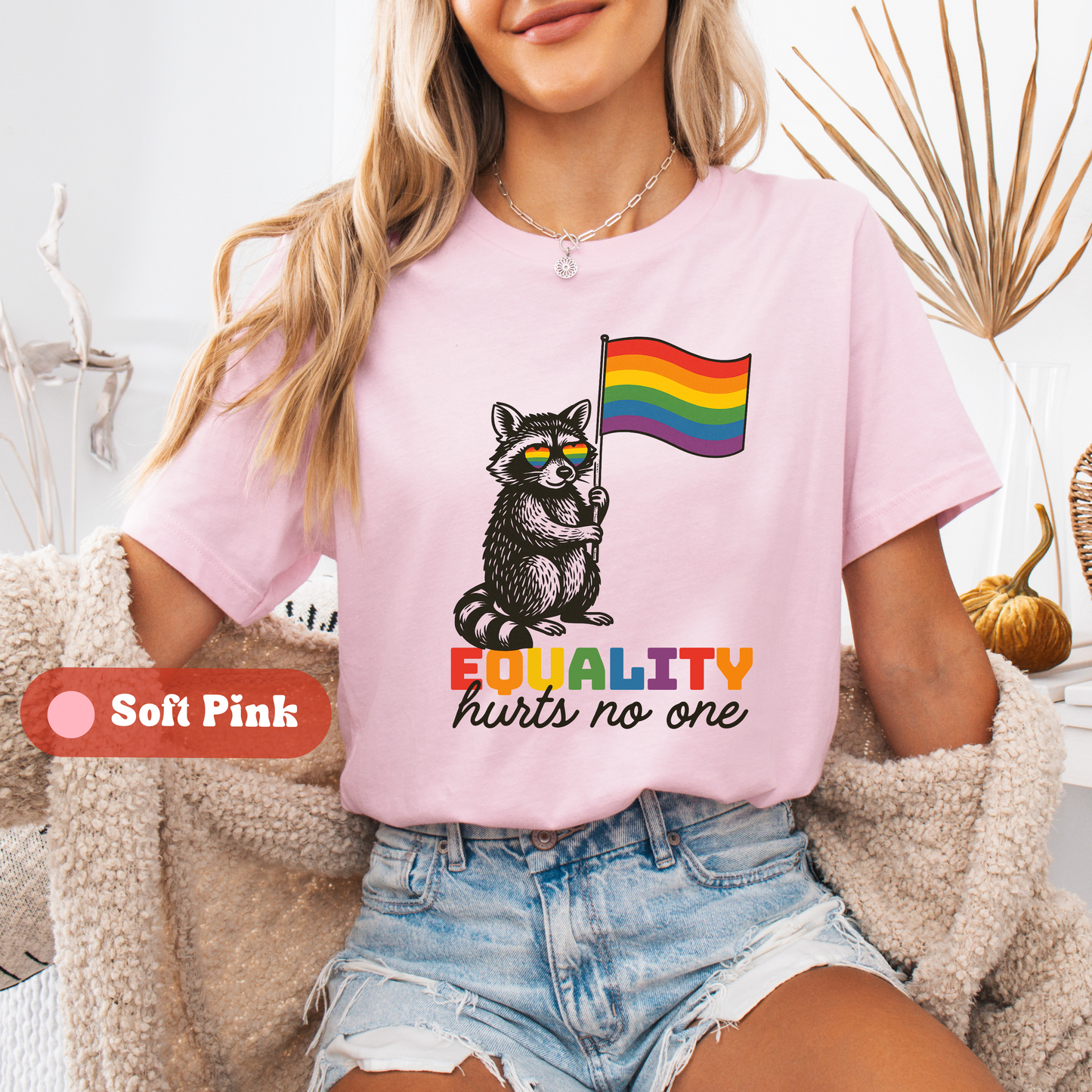 A woman wearing a soft pink t-shirt with a rainbow pride raccoon design and the message βEQUALITY hurts no one.β A label notes the color as Soft Pink.
