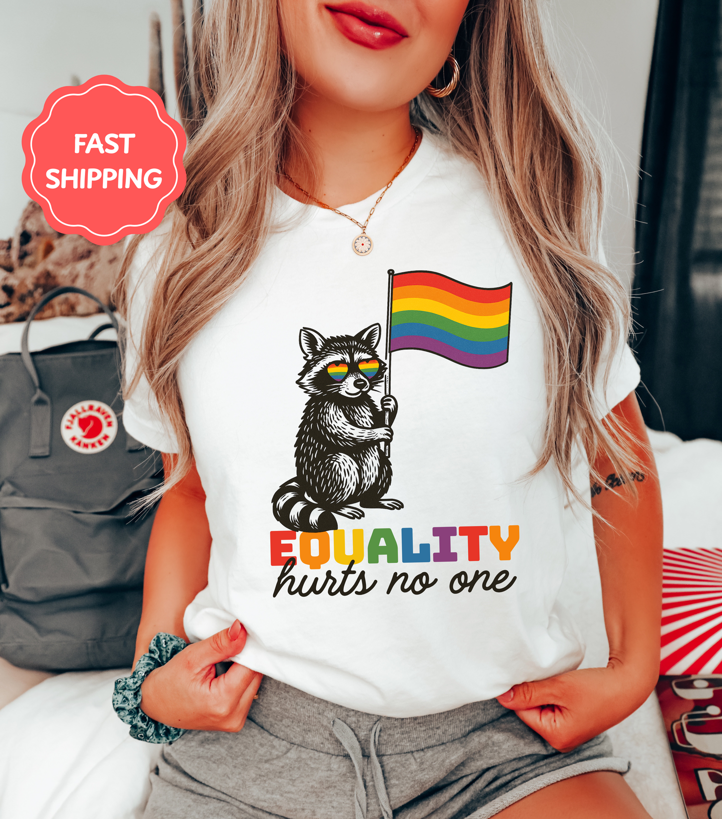 A woman wearing a white t-shirt featuring a black and white raccoon holding a rainbow flag with the text βEQUALITY hurts no one.β A red βFast Shippingβ badge appears in the corner.